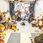 A children's play room, full of colourful toys, two toddlers are playing with a toy house, watched over by their parents