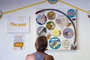 A person at an exhibition, looking at a collaborative collage artwork labelled "The Past"