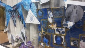 A photograph of a window, decorated with blue fabric bunting, some of it hand dyed, and blue-patterned paper bunting decorated with black and white images of people. A board covered with black and white pictures of people, which ahve been painted over with blue and yellow paint. There is a bunch of blue flowers on the windowsill.
