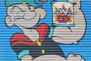 A photograph of a closed shop shutter, with a street art image of Pop-Eye the sailor, holding a can of spinach labelled "Power Up"