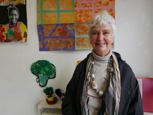 Mo Gallaccio standing in front of a wall displaying bright and colourful children's paintings 