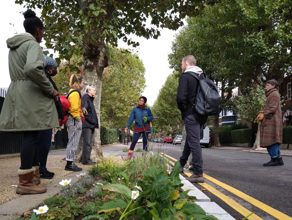 A group of people on a guided walk, standing around a pavement flower bed, listening to a speaker.
