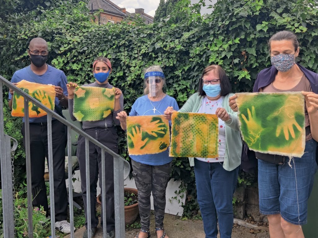 Five people standing in The Mill garden holding up art works they've made, using brigh greens and yellows to create silhouettes on cloth
