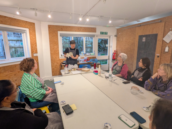 A room with windows and a door along one wall, open to a garden. In the centre of the room is a large table, with 3 chairs spaced to allow for 2 metres social distancing